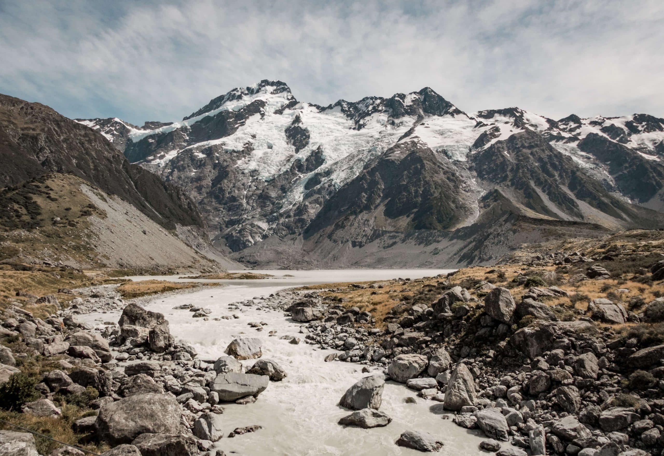 A rocky river bed flowing from a lake, in front of a snow-capped mountain range.
